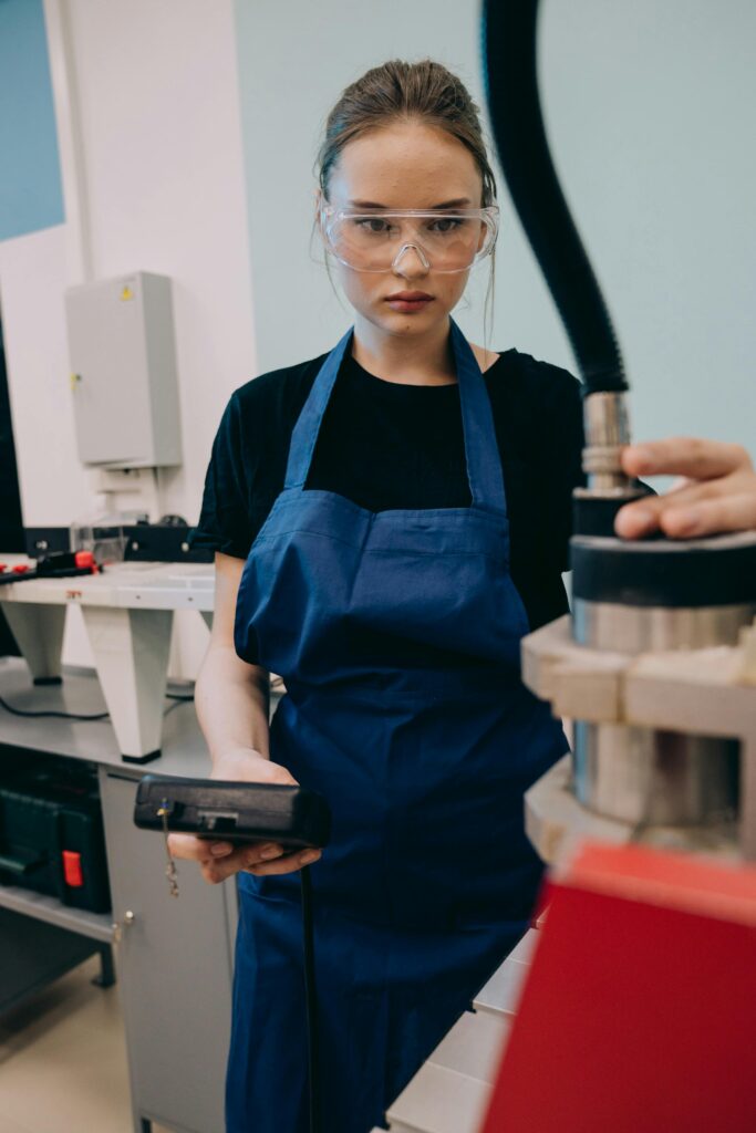 Young woman in workshop using machinery, wearing protective goggles and blue apron.